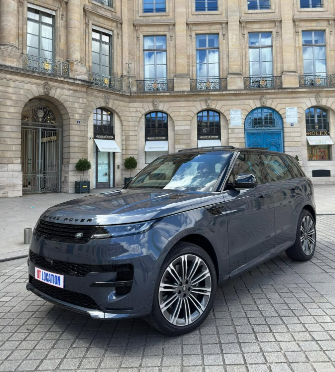 Un SUV gris foncé est garé dans une rue pavée, devant un bâtiment en pierre orné.