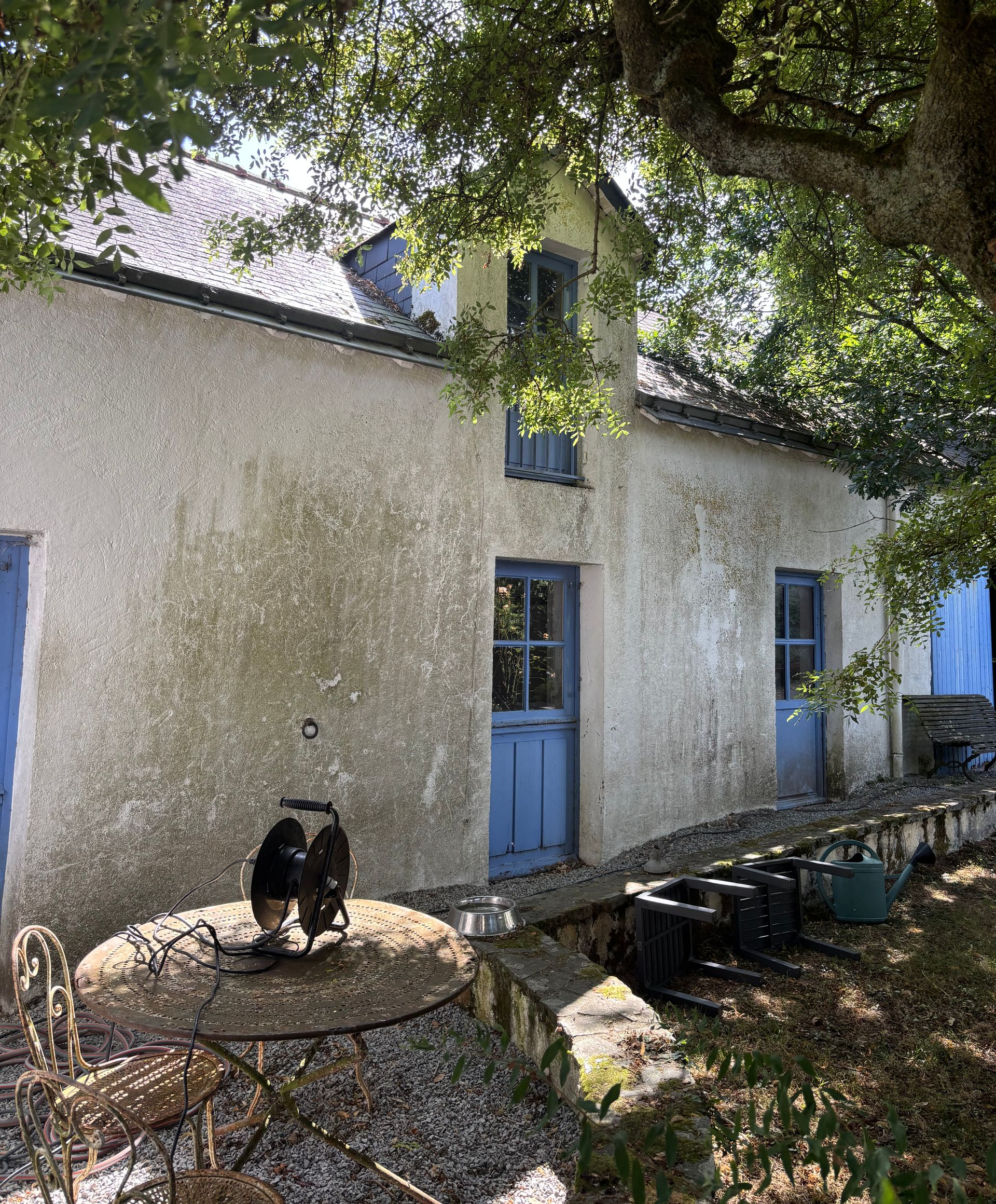 Maisonnette en pierre aux portes et fenêtres bleues, sous un grand arbre, avec un chat noir assis sur une table devant.