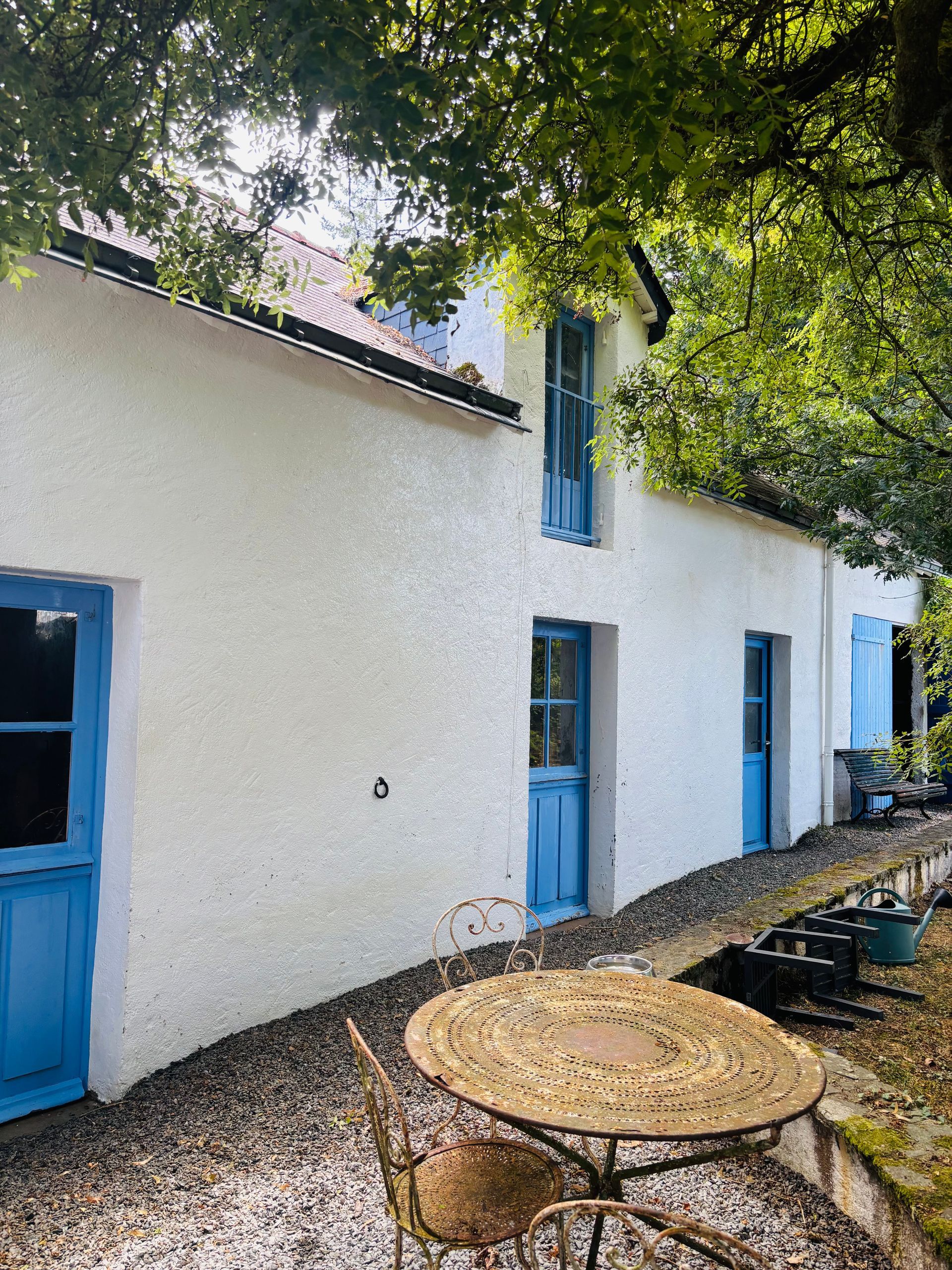 Une maison blanche aux portes et fenêtres bleues, avec une table ronde en fer forgé et des chaises sur une terrasse en gravier sous un grand arbre.