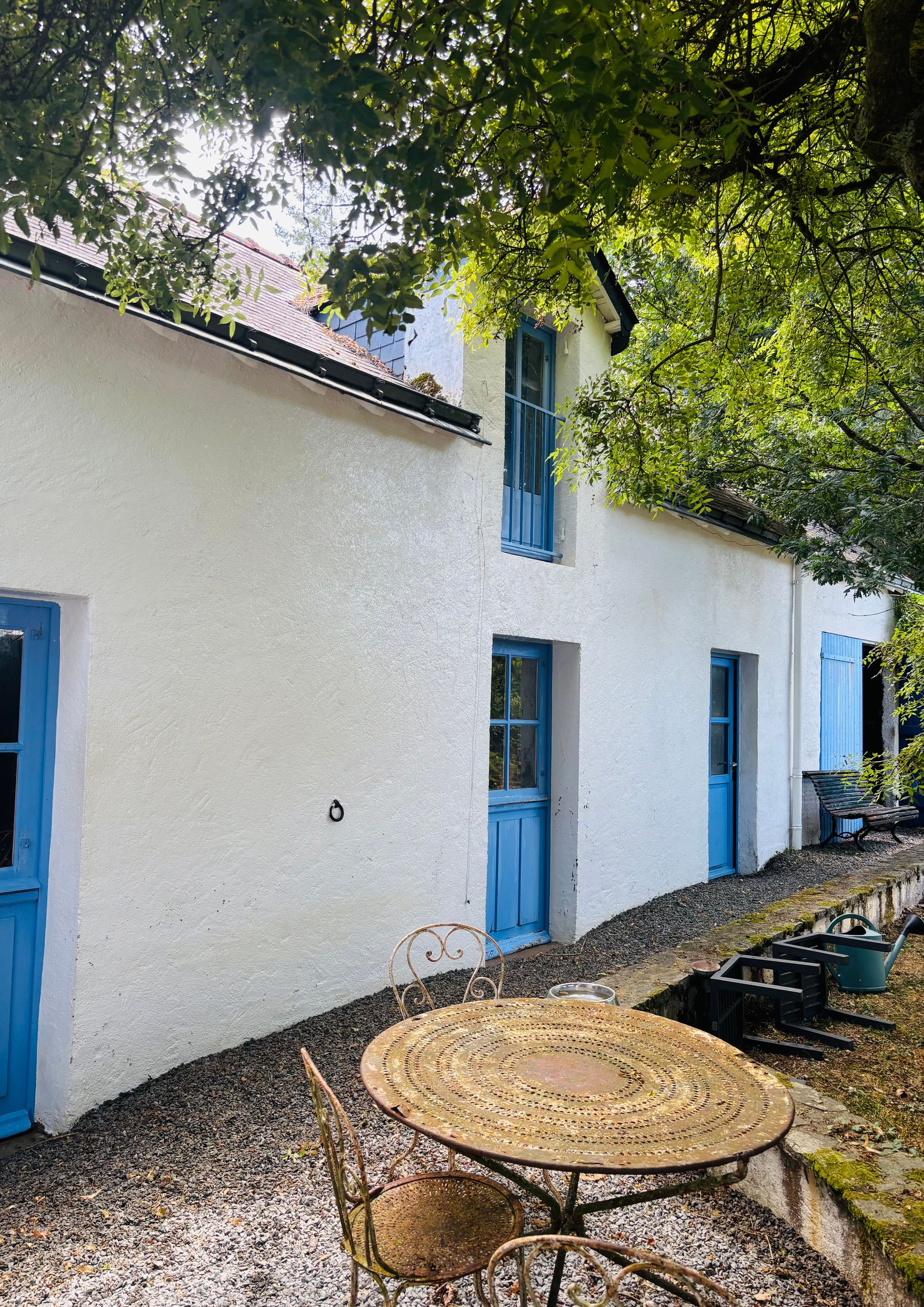 Maison blanche avec portes et fenêtres bleues, jouxtant un patio arboré avec tables et chaises en métal.