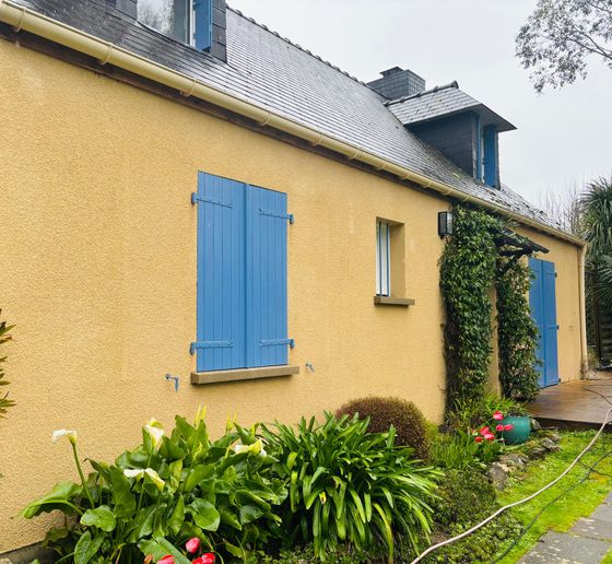 Une maison jaune au toit d'ardoise, aux volets bleus et devant laquelle poussent des plantes d'un vert éclatant.