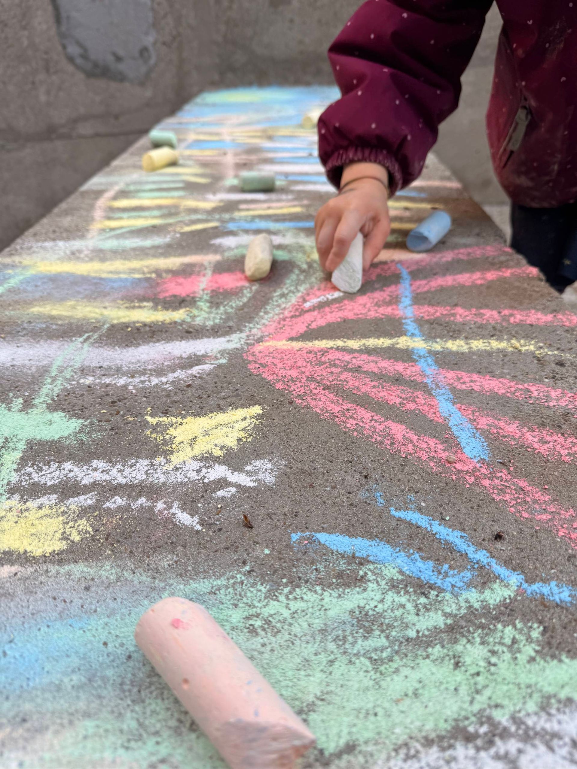 Ein Kind zeichnet bunte Kreidelinien auf den Bürgersteig; im Vordergrund ist ein rosa Kreidestift zu sehen.