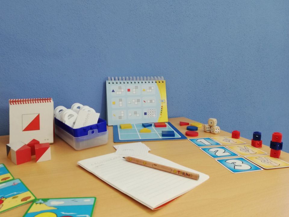 Schreibtisch mit Spielen, Bauklötzen, Notizblock, Bleistift und Würfeln, möglicherweise für eine Lerntherapie in einem Raum mit blauen Wänden.
