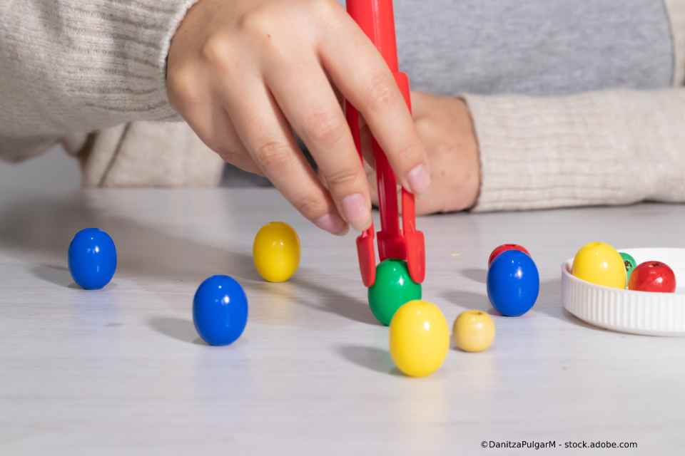 Eine Person nimmt mit einer roten Zange ein grünes, eiförmiges Spielzeug von einem Tisch mit anderen farbigen Spielzeugen.