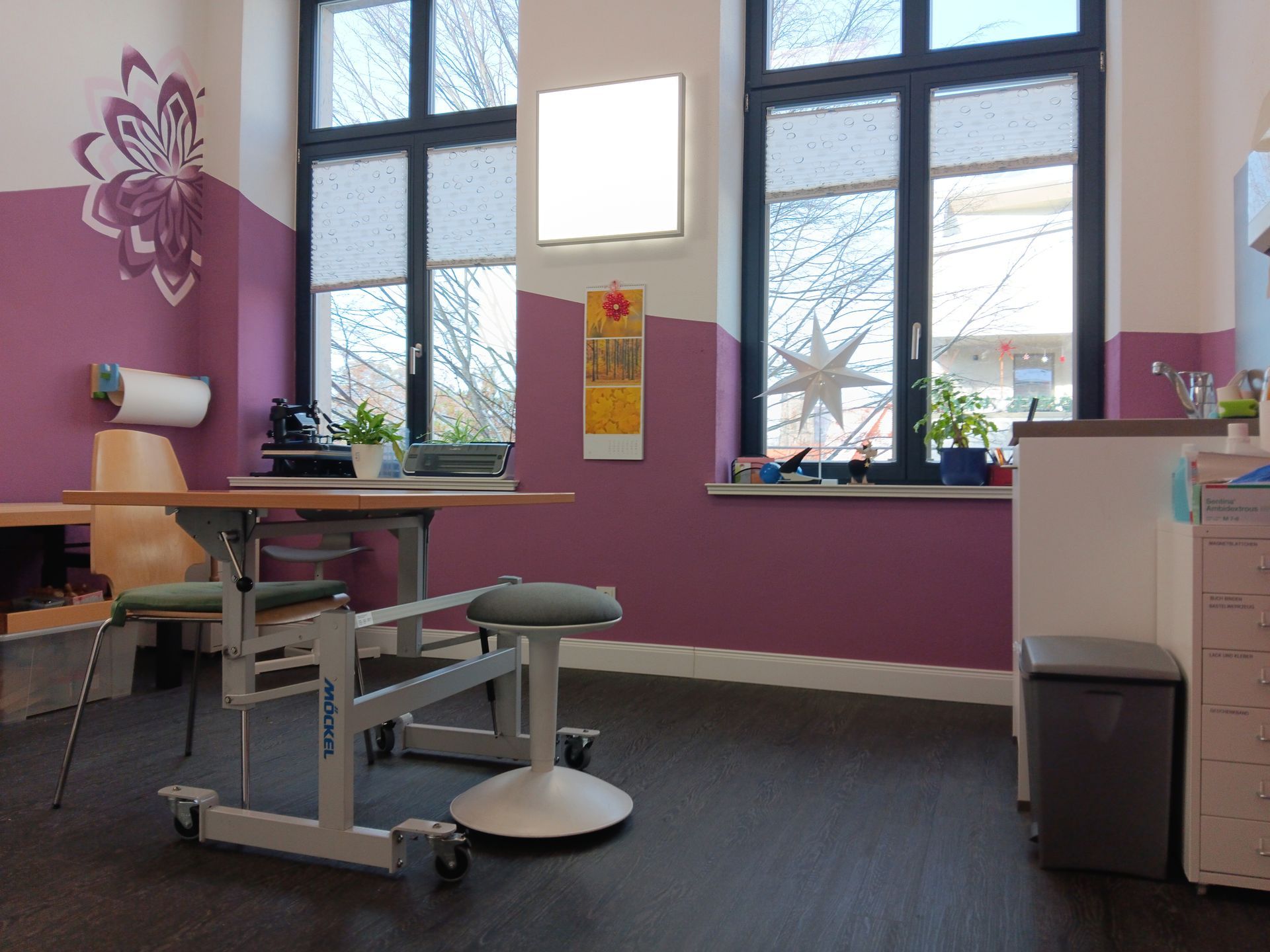 Zimmer mit einem Schreibtisch, zwei Fenstern und violetten Wänden. Ein Blumenmuster schmückt eine Wand.