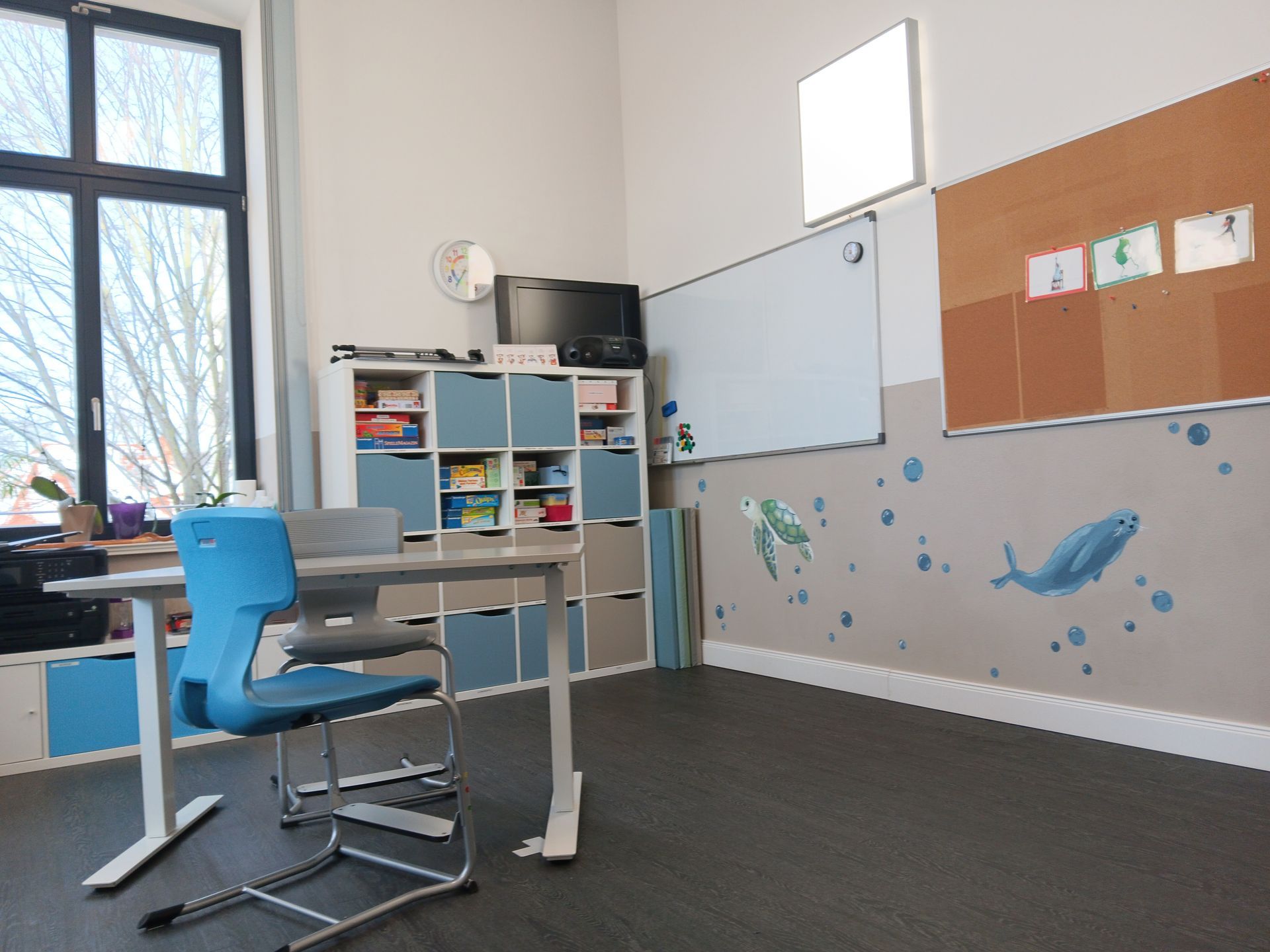 Klassenzimmer mit Schreibtisch, Stuhl, Bücherregal, Whiteboard, Fernseher, Pinnwand und Wandkunst mit Meeresmotiven.