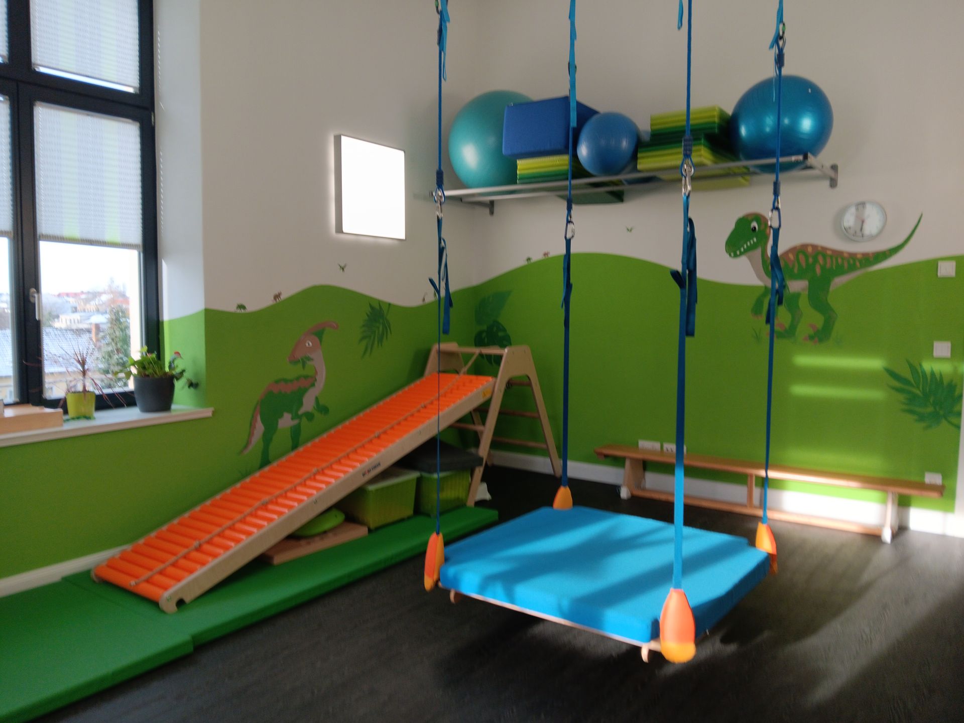 Spielzimmer mit Schaukel, Rutsche und Dinosaurier-Wandbild. Grüne und blaue Akzente.