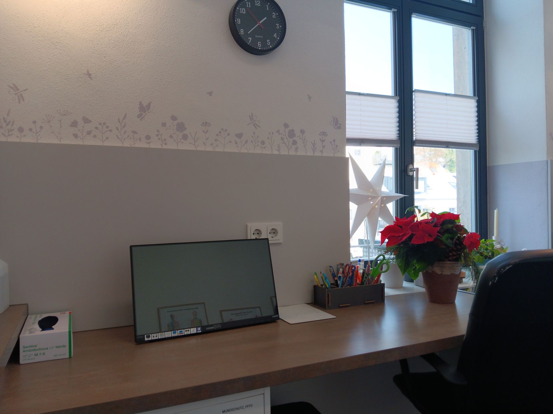 Schreibtisch mit Computermonitor, Uhr und Fenster mit Blumen; Büroumgebung.