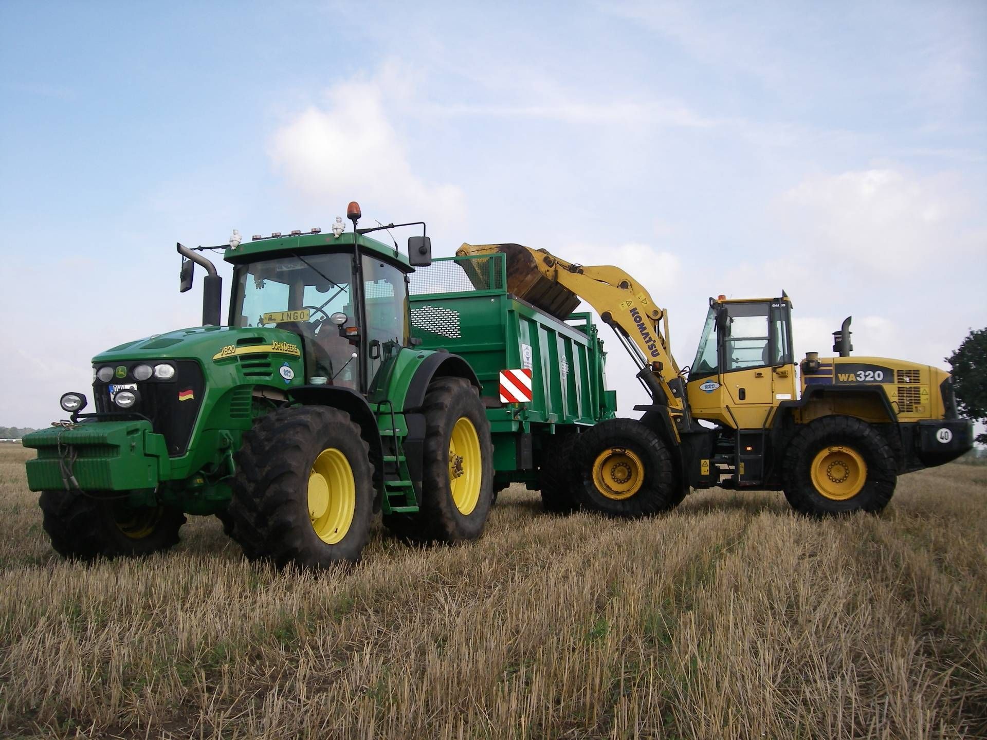 Ein grüner John Deere Traktor steht neben einem gelben Bagger auf einem Feld