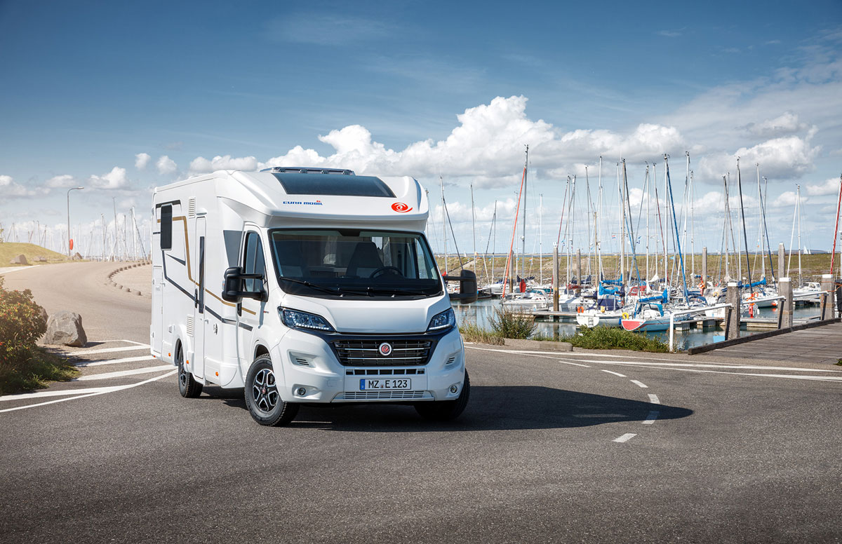 Ein weißes Wohnmobil ist am Straßenrand neben einem Yachthafen geparkt.