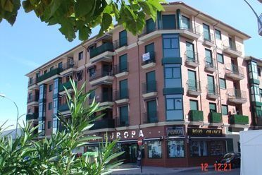 Edificio de apartamentos de ladrillo con balcones verdes y locales comerciales en una esquina soleada.