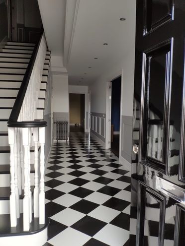 Pasillo con suelo de damero blanco y negro, escalera, barandillas blancas y puerta principal oscura.