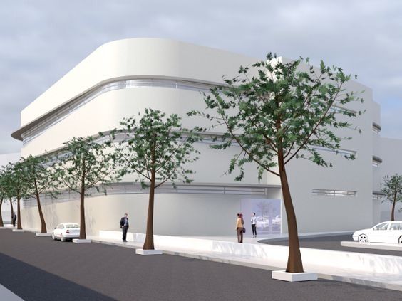 Edificio blanco moderno en una calle de la ciudad con árboles, peatones y coches bajo un cielo nublado.