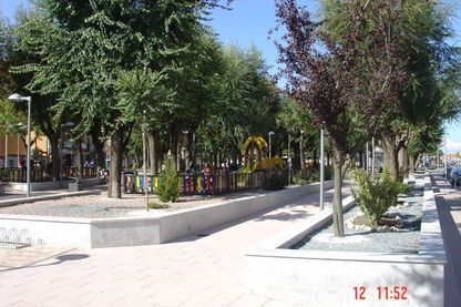 Plaza urbana arbolada con jardineras elevadas, sendero pavimentado y bancos a plena luz del día.