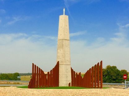 Monumento alto de hormigón con paneles metálicos curvos de color óxido en una rotonda cubierta de hierba bajo un cielo azul.