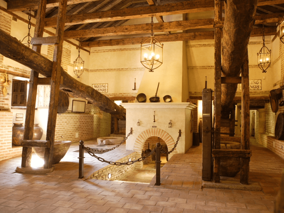Habitación rústica histórica con vigas de madera, chimenea central y faroles colgantes bajo una iluminación cálida.