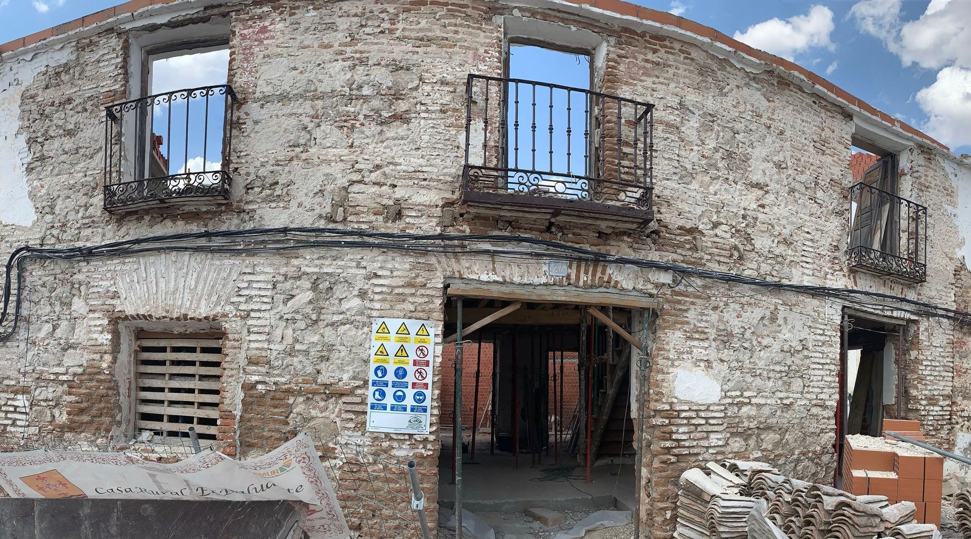 Edificio de piedra desgastada en proceso de renovación, con ventanas enrejadas, una puerta abierta y materiales de construcción en el exterior.