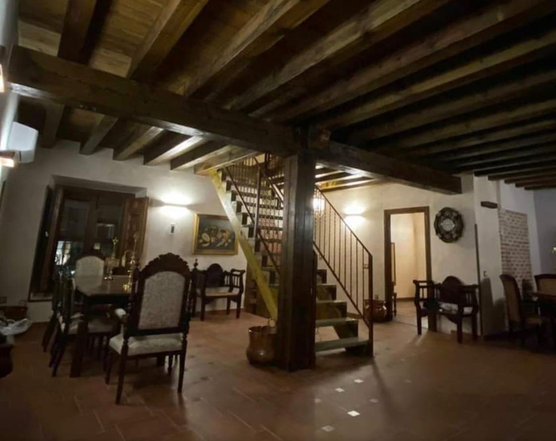 Acogedor comedor rústico con vigas de madera, escalera, mesas y sillas bajo una iluminación cálida.