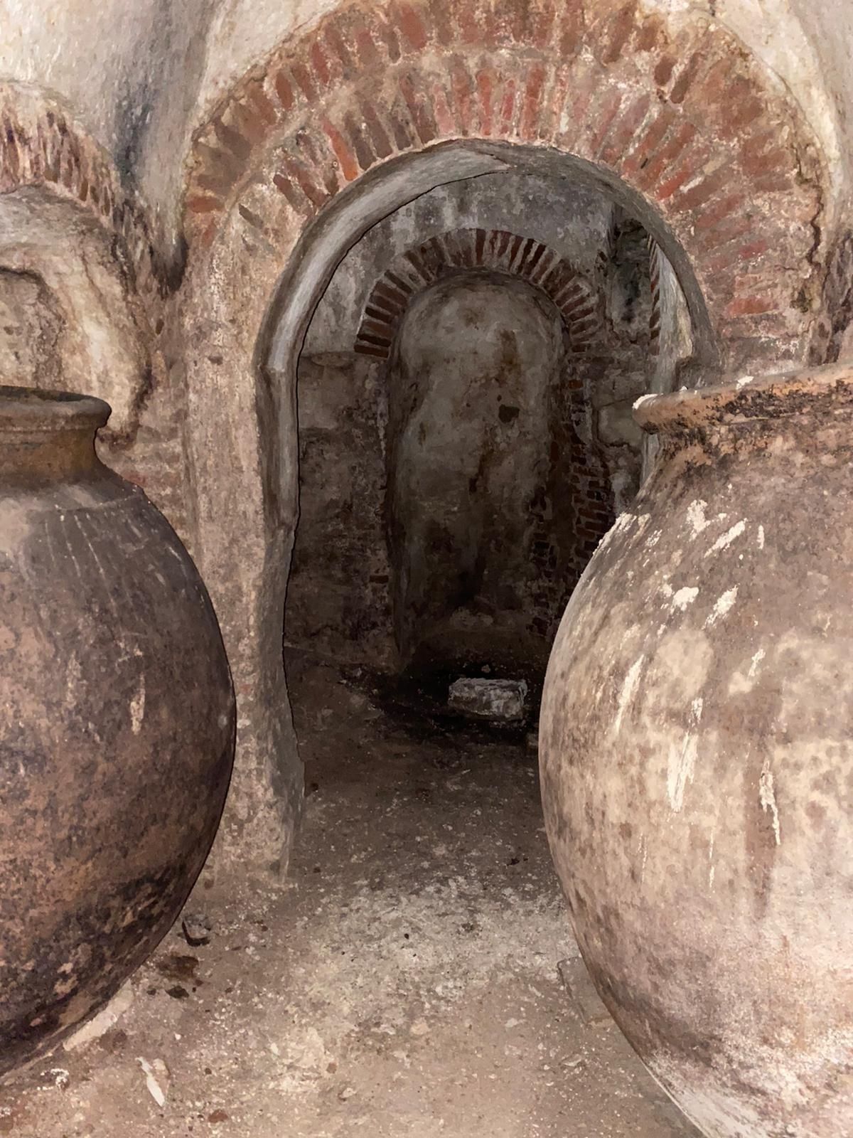 Antigua bodega de ladrillo con grandes tinajas de barro flanqueando una estrecha puerta arqueada.