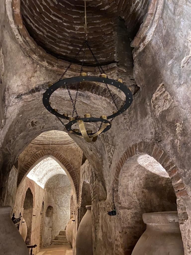 Interior de piedra antigua con techos abovedados y una lámpara de araña circular de metal que cuelga sobre un pasillo.
