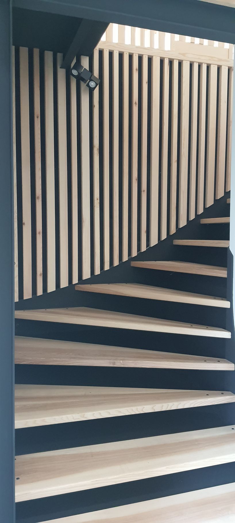 Escalier en bois avec paroi à lattes verticales. Marches et contremarches claires avec supports en acier noir.