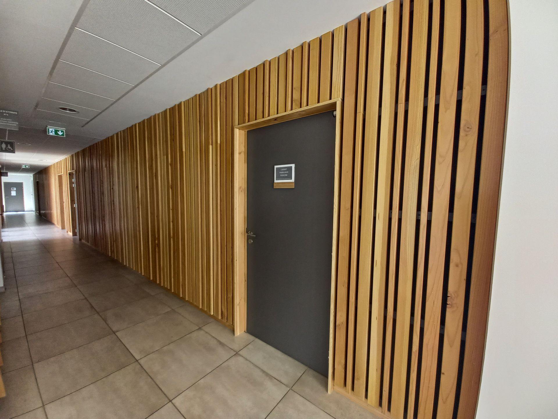 Couloir avec mur en lattes de bois, porte grise, sol en carrelage et éclairage encastré.