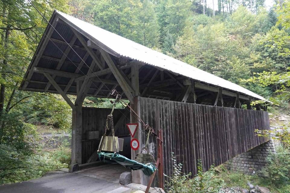 Überdachte Holzbrücke mit Giebeldach, die eine Straße in einem Waldgebiet überspannt.