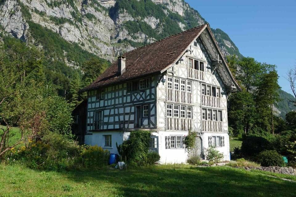 Fachwerkhaus mit braunem Dach und weissen Wänden, vor einer Bergkulisse und grünem Gras.