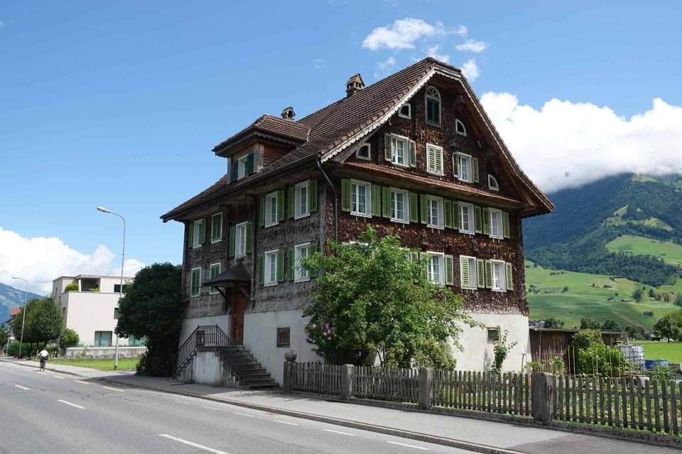 Braunes Holzhaus mit grünen Fensterläden und Giebeldach vor einer Bergkulisse und blauem Himmel.