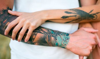 Brazos entrelazados, luciendo coloridos tatuajes. Se ve una camisa blanca, lo que sugiere una pareja abrazándose.
