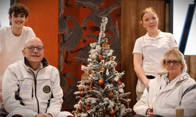 Quatre personnes posent près d'un sapin de Noël décoré. Deux sont assises, deux sont debout, toutes vêtues de vestes blanches. Fond mural orange.