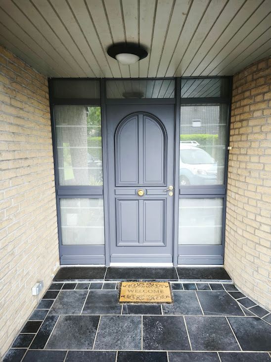 Porte d'entrée grise avec imposte, sous un porche couvert en briques et sol carrelé foncé.
