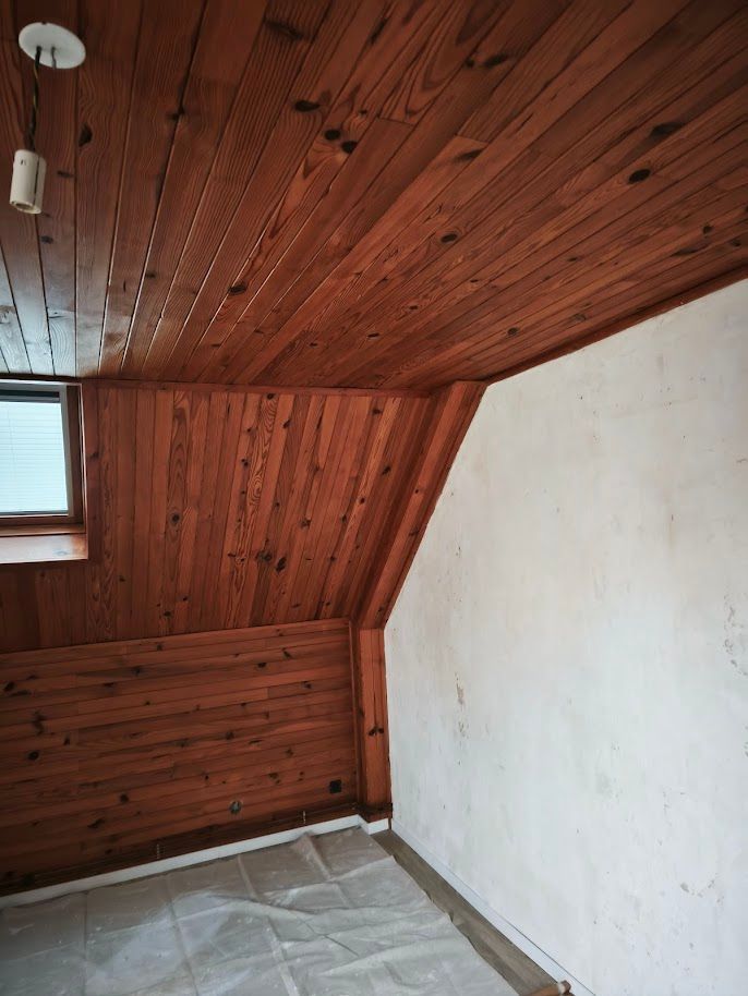 Plafond et murs lambrissés, petite fenêtre, mur blanc, sol recouvert.