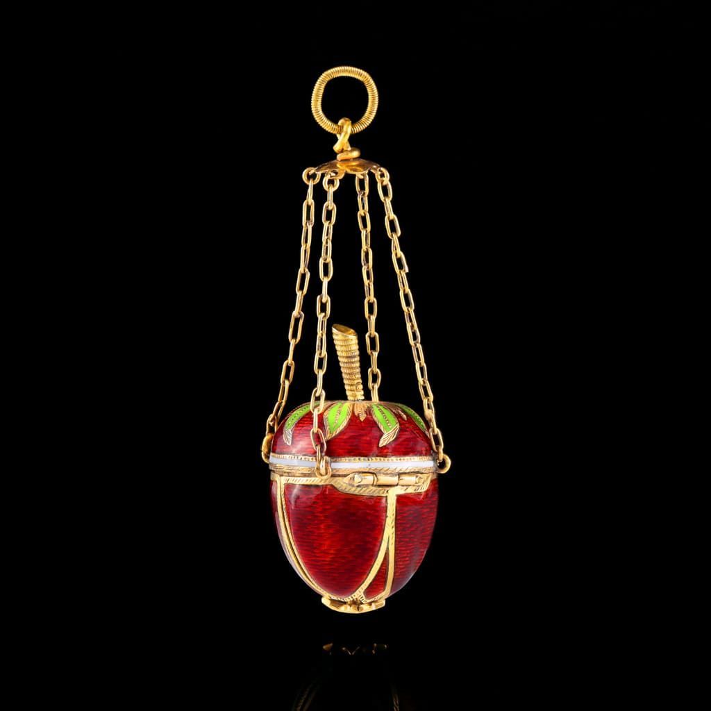 Un petit pendentif en forme de pomme rouge émaillée, dans une cage en or, suspendu par quatre délicates chaînes en or sur fond noir.