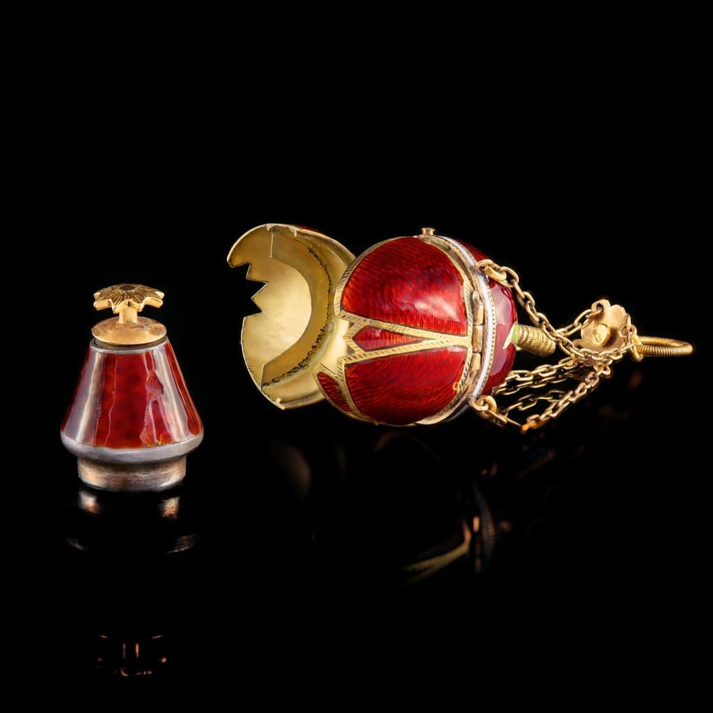 Un petit pendentif en forme d'œuf Fabergé ouvert, émaillé rouge, avec des filigranes d'or et des chaînes attachées, sur fond noir.