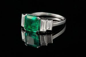 Une bague en argent ornée d'une pierre précieuse verte centrale taille émeraude, flanquée de deux diamants baguette sur fond noir.