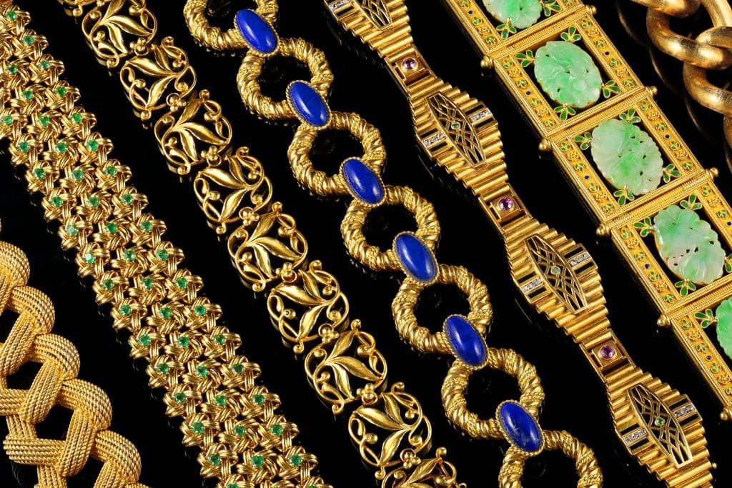 Une collection de bracelets en or variés ornés de pierres précieuses, notamment du lapis-lazuli bleu et du jade vert, sur une base noire.
