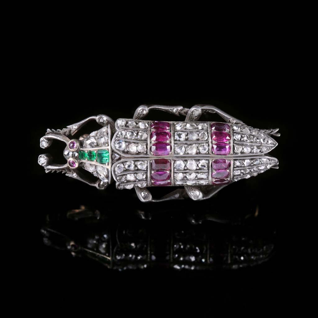 Broche scarabée ornée de diamants, de rubis rouges et d'émeraudes vertes sur fond noir réfléchissant.