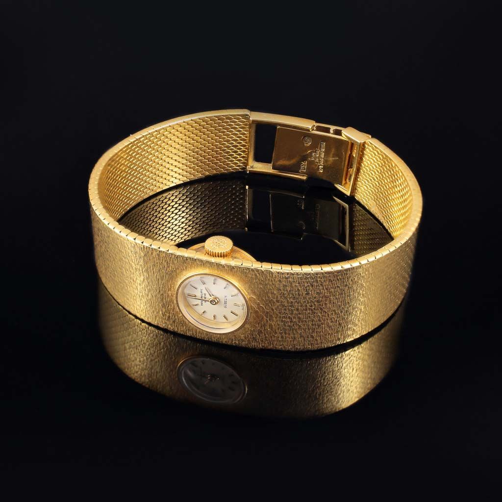 Une montre en or texturé avec un cadran blanc circulaire, montée sur un large bracelet manchette en or tressé sur fond noir.