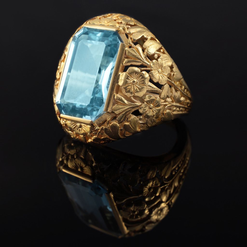 Bague en or ornée d'une grande pierre précieuse rectangulaire bleu clair sertie sur un anneau finement gravé de motifs floraux et de feuilles.