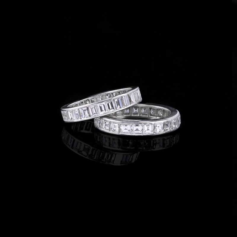 Deux anneaux en platine sertis de diamants taille émeraude, posés en rangée sur une surface noire réfléchissante.