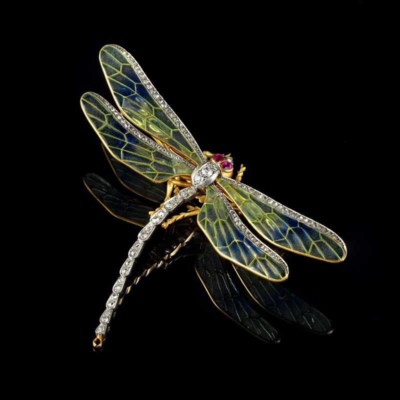 Broche libellule aux ailes translucides bleues et vertes, au corps incrusté de diamants et aux yeux en rubis sur fond noir.