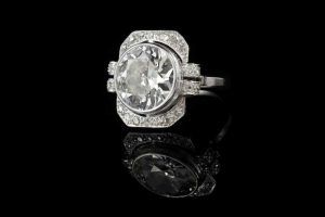 Une bague en diamant de style vintage avec une pierre centrale ronde sertie dans de l'argent, se reflétant sur une surface noire brillante.