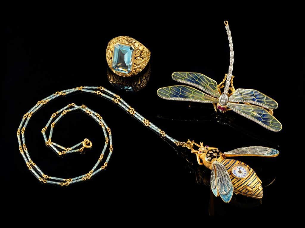 Bijoux en or sur fond noir : une broche libellule, un pendentif abeille sur une chaîne assortie et une bague rectangulaire bleue.