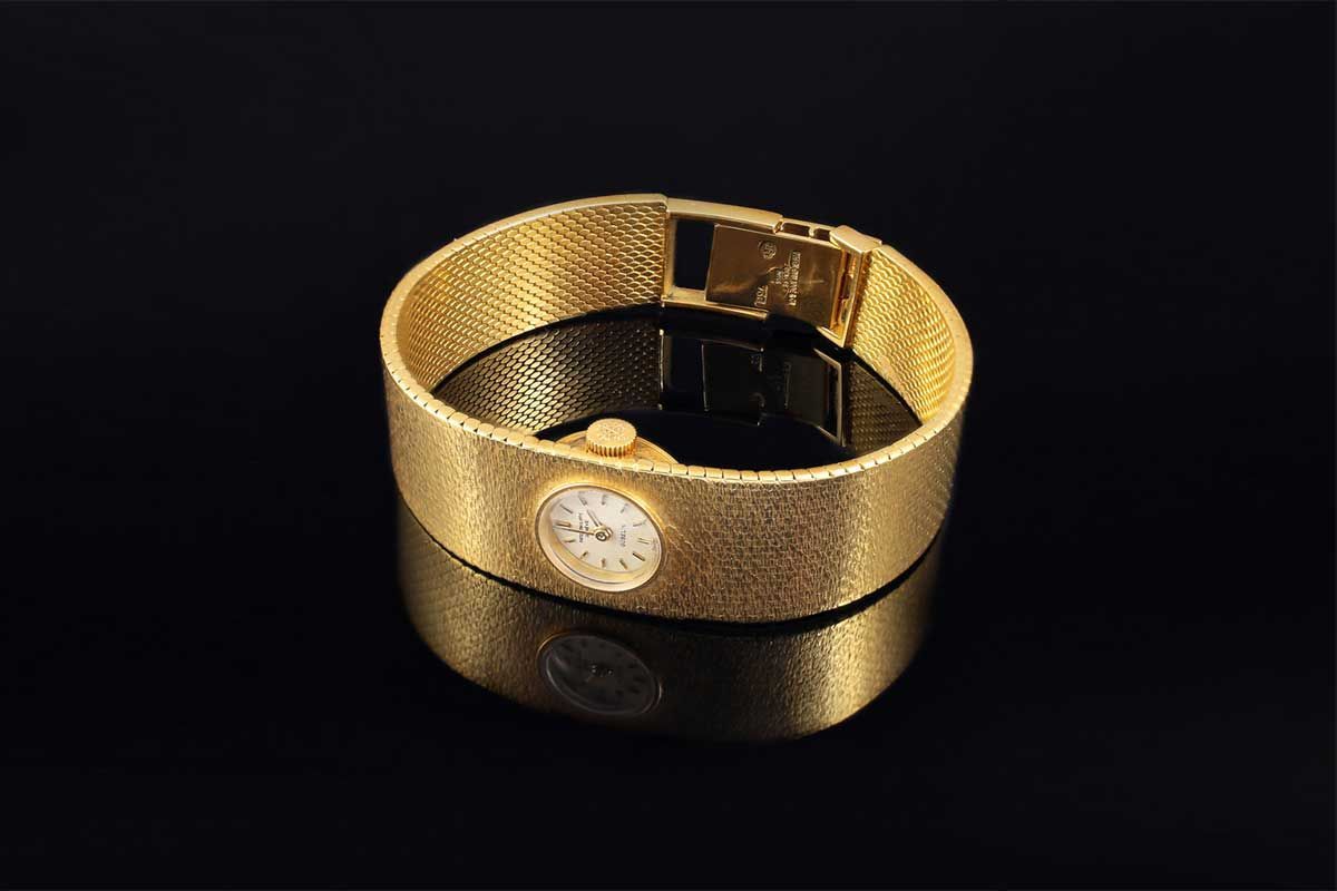 Une montre-bracelet en or à la finition texturée et au cadran blanc circulaire sur fond sombre et réfléchissant.