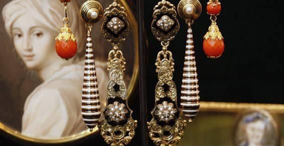 Deux longues boucles d'oreilles pendantes ornées de perles rouges, de cônes rayés noirs et blancs et de filigranes dorés, sur fond de tableaux.