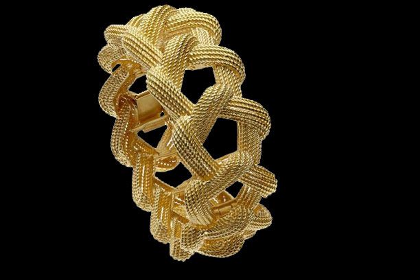 Un large bracelet en or texturé, orné d'un motif complexe tissé en forme de nœud sur fond noir uni.