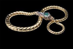 Bracelet serpent en or avec un corps articulé et enroulé, orné d'une tête en émeraude et diamant sur fond noir.