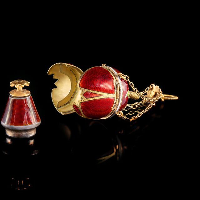 Un pendentif en forme d'œuf ouvert, émaillé rouge et doré, avec un embout amovible assorti, sur une surface noire réfléchissante.