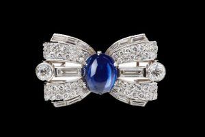 Broche en forme de nœud avec un saphir cabochon bleu central flanqué de diamants sur fond noir.